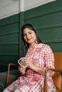Positive smiling Indian lady in stylish traditional wear sitting on chair against green wall with cup of hot drink and looking at camera